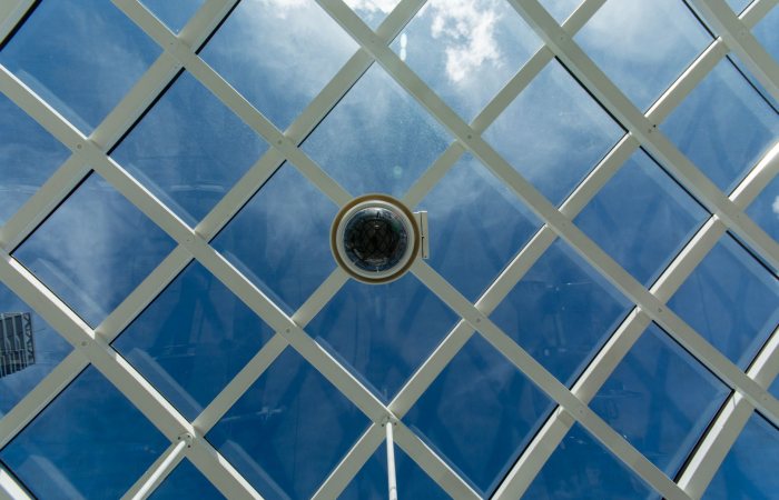 Surveillance camera mounted on a modern glass ceiling with sky reflections.