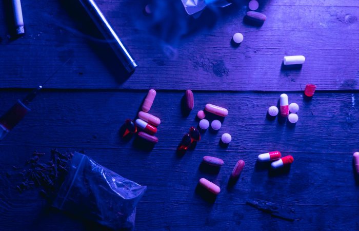 A flat lay of various drugs including pills, capsules, and a syringe on a wooden surface under moody blue lighting.